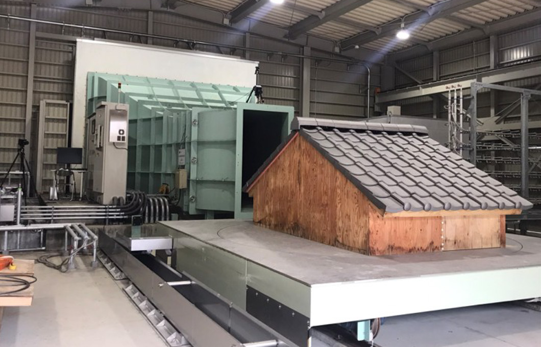 大型の送風装置で、家の模型に強い風を当てて影響を調べる実験の様子。装置の前に屋根付きの家の模型が置かれている。：写真