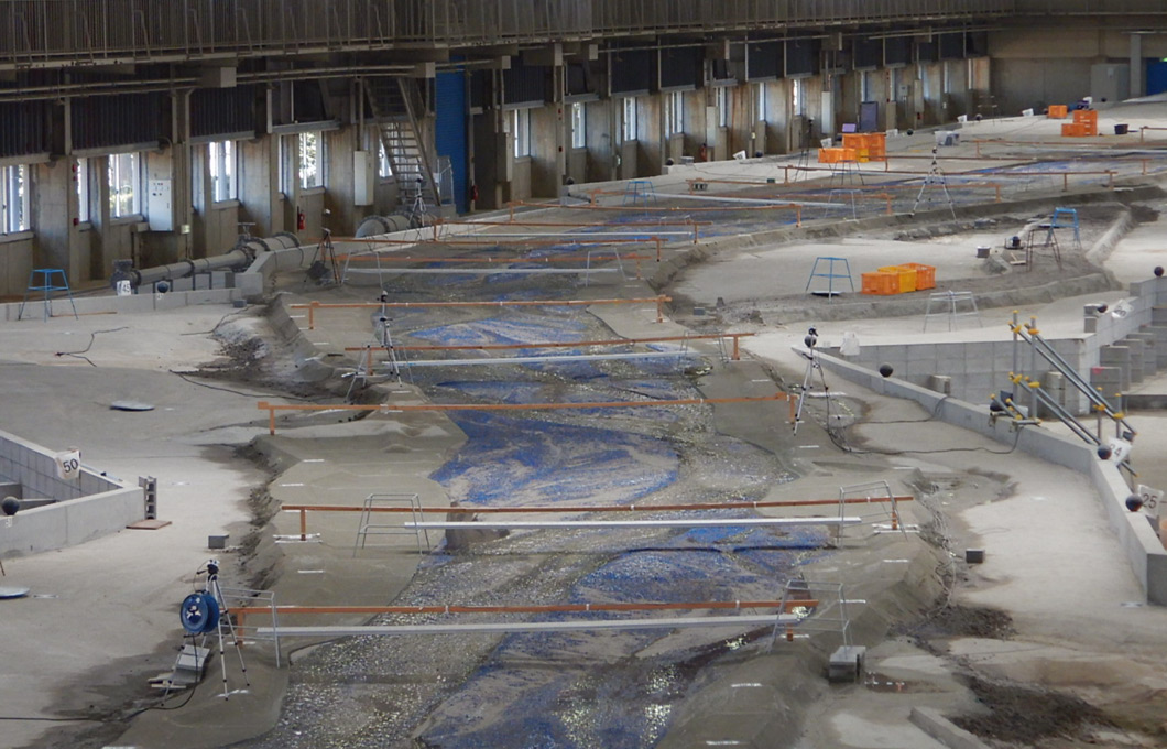 水理模型実験施設の様子。屋内の広い実験場に、本物の川を小さく再現した模型があり、水を流して川の形や流れ、土砂の動きを調べている。：写真