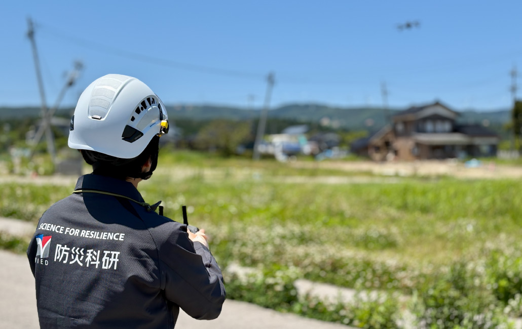 ヘルメットを着けた防災科研の職員が、野外でドローンを操作している写真。災害の被害状況を空から調べるためにドローンが使われている。：写真